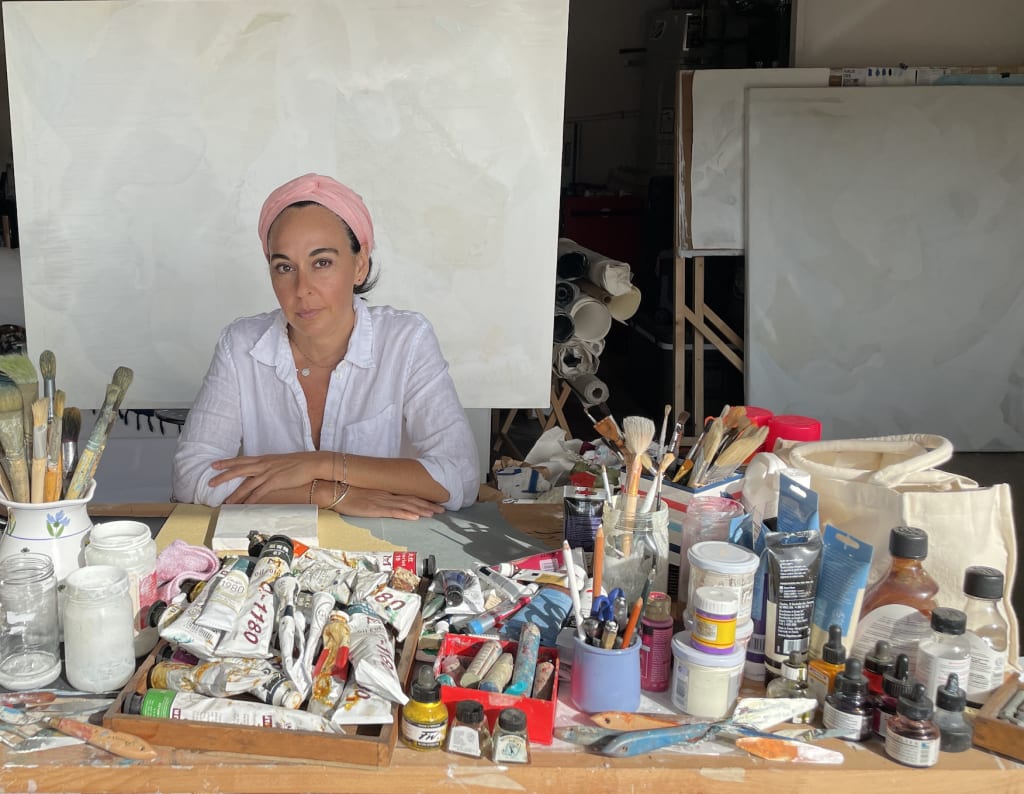 Joanna Cutri, a painter, sits at a table covered with paints, brushes, and other artistic supplies